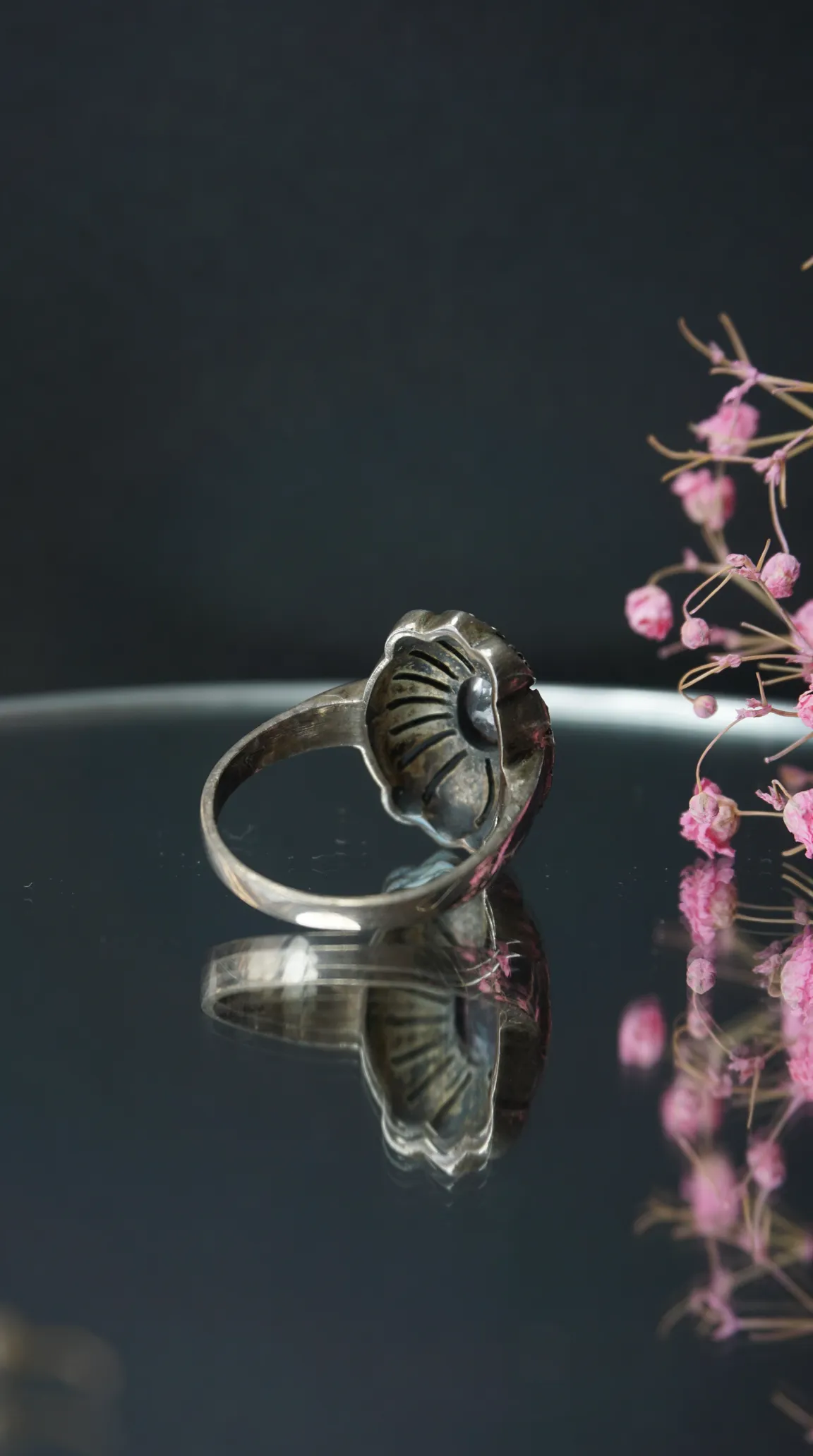 Vintage silver ring with intricate scalloped design on a dark reflective surface, with soft pink dried flowers.