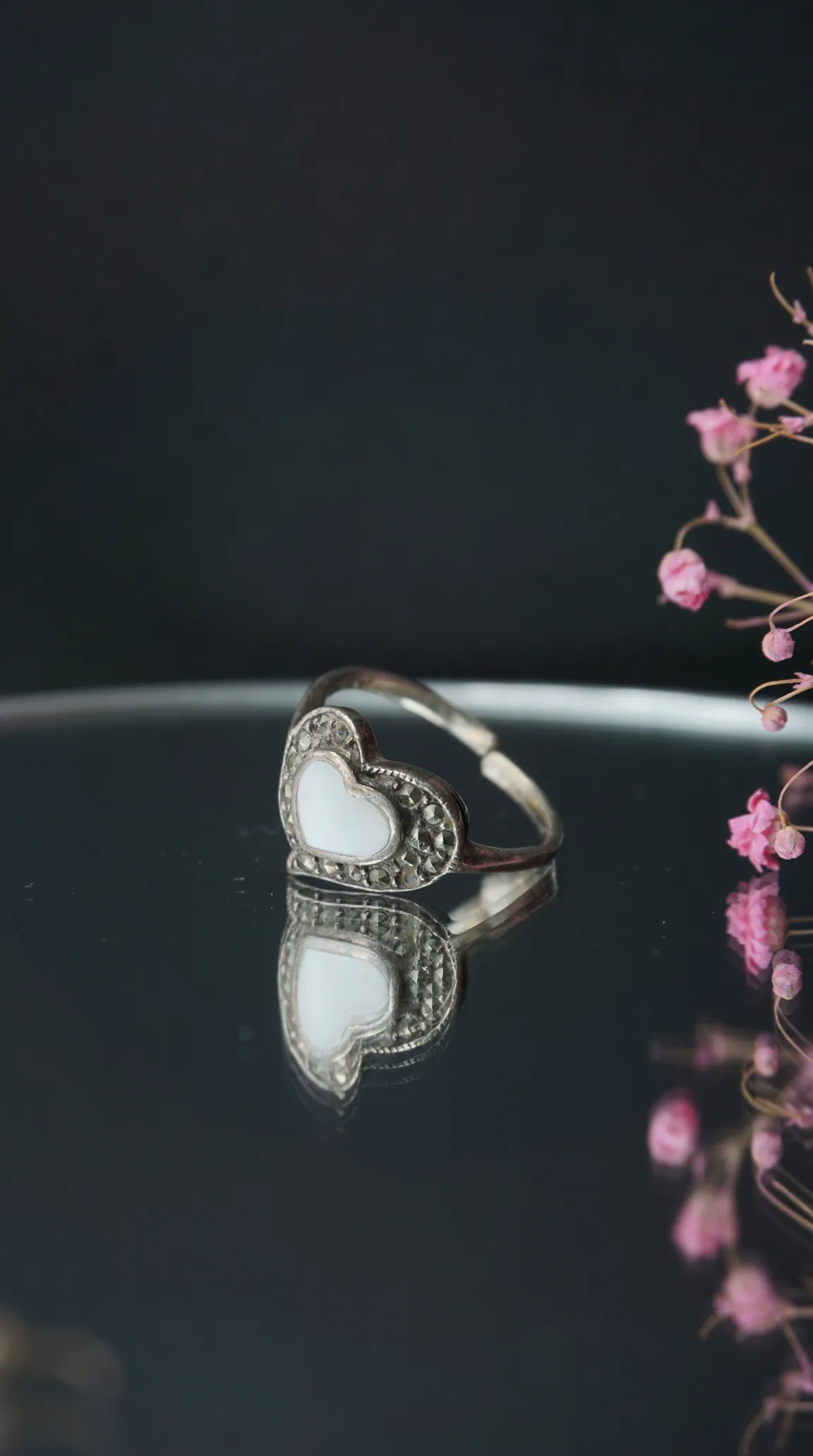 Vintage sterling silver heart ring with mother of pearl inlay and marcasite accents on a reflective surface, with soft pink flowers.