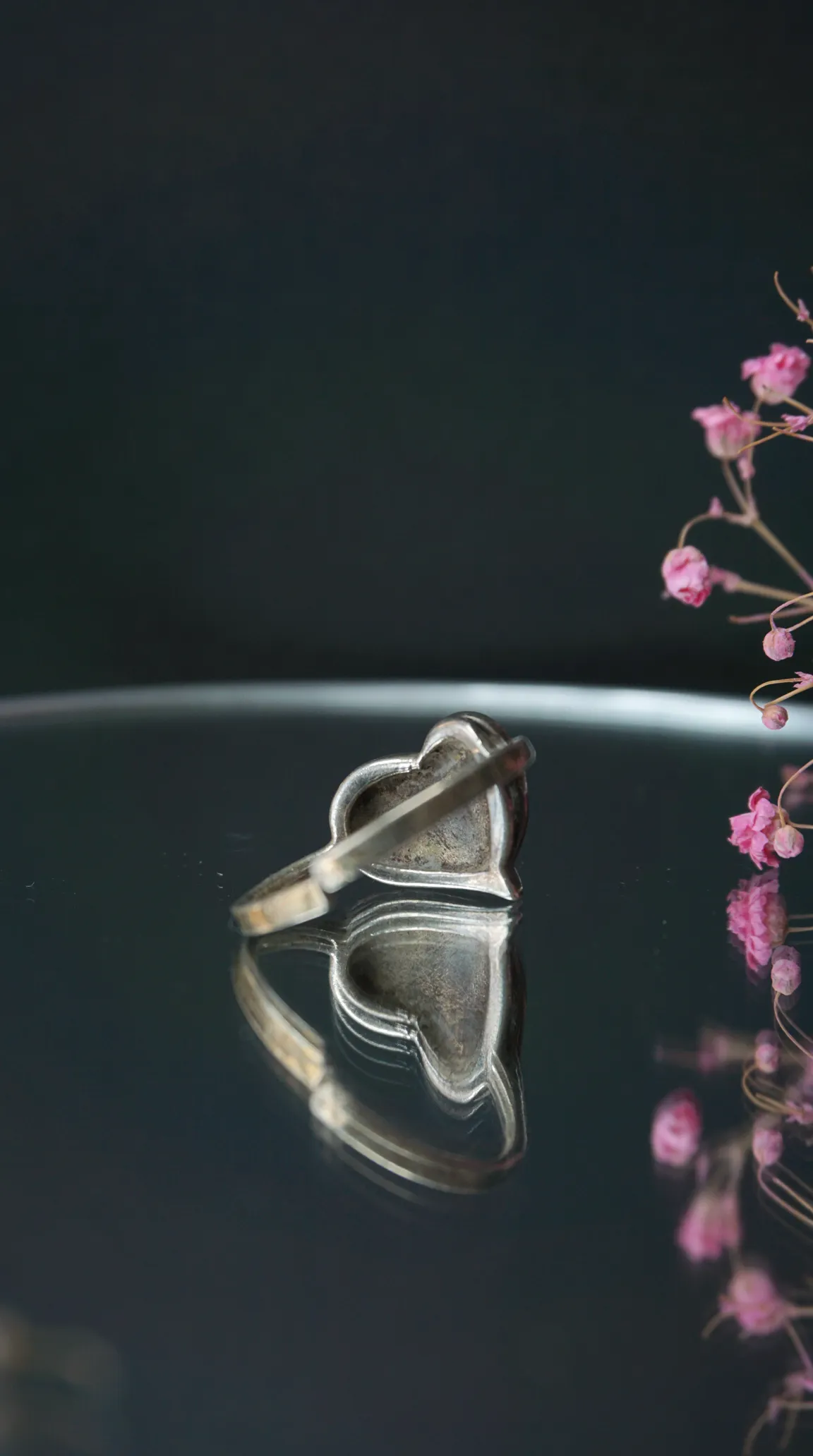 Vintage heart-shaped silver ring on dark reflective surface with soft pink flowers.