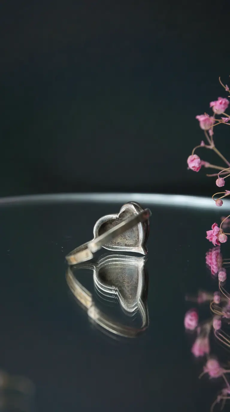 Vintage heart-shaped silver ring on dark reflective surface with soft pink flowers.