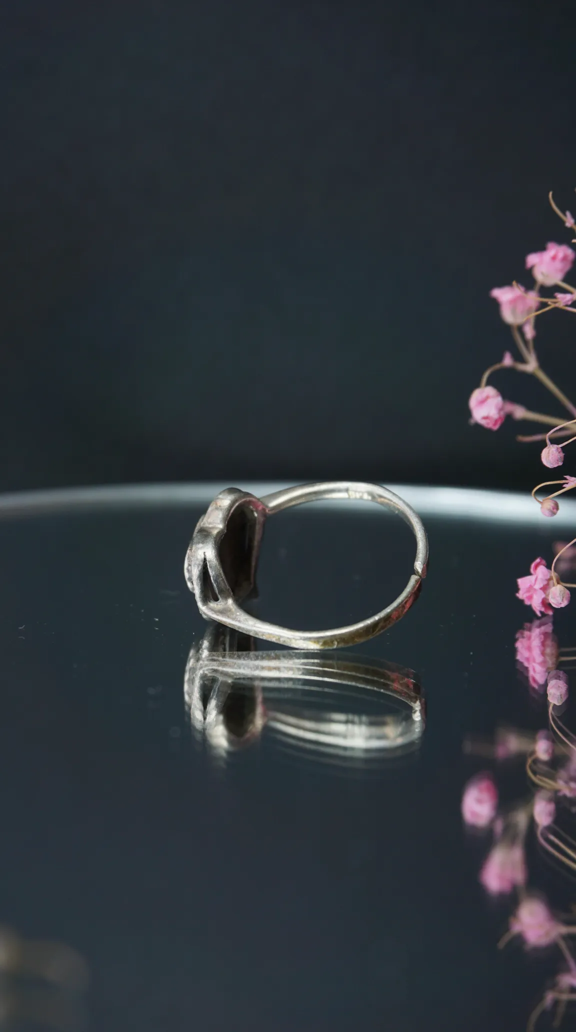 Vintage silver ring with ornate dark gemstone setting, reflecting on a polished surface with pink dried flowers.