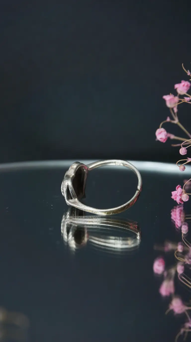 Vintage silver ring with ornate dark gemstone setting, reflecting on a polished surface with pink dried flowers.
